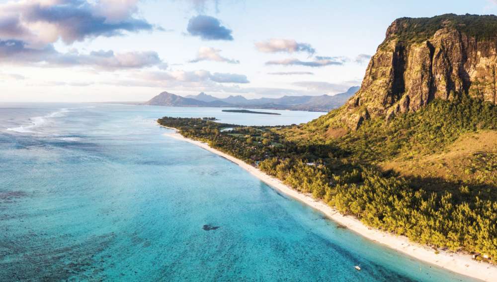 A distant image of a long beach in Mauritius. There are mountains in the distance.